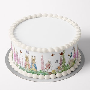 May include: A round cake with white frosting and a decorative border. The cake's side features a whimsical illustration of rabbits, butterflies, and flowers in a garden setting. The cake sits on a silver-coloured cake board.