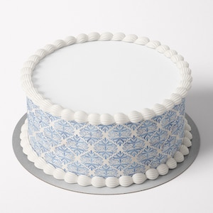 May include: A round cake with white frosting on top and around the base. The sides of the cake are decorated with a blue and white floral pattern. The cake sits on a silver-colored cardboard base.