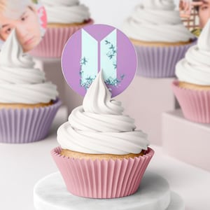 Puede incluir: Cupcakes con glaseado blanco en moldes rosas y morados. Cada cupcake tiene un adorno decorativo con un logotipo y detalles florales. Los cupcakes están sobre un soporte de mármol blanco.