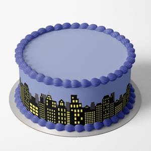 Puede incluir: Un pastel redondo con una parte superior de glaseado azul claro y un borde de glaseado azul oscuro. El pastel presenta un diseño de paisaje urbano negro y amarillo alrededor de los lados, con edificios y ventanas iluminadas. El pastel se encuentra sobre una base de pastel plateada.
