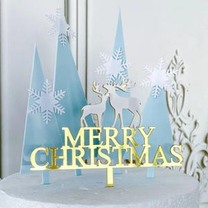 Könnte beinhalten: Ein Tortenaufsatz mit Winterthema. Er zeigt hellblaue dreieckige Bäume, weiße Schneeflocken und zwei weiße Rentiere. Die Worte "MERRY CHRISTMAS" sind in Gold gehalten. Zum Aufstellen auf einem Kuchen.