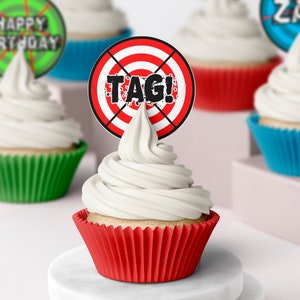 May include: Red and white target cupcake topper with the text "TAG!" in black. The topper is on a cupcake with white frosting and a red cupcake liner. The cupcake is on a white marble cake stand.