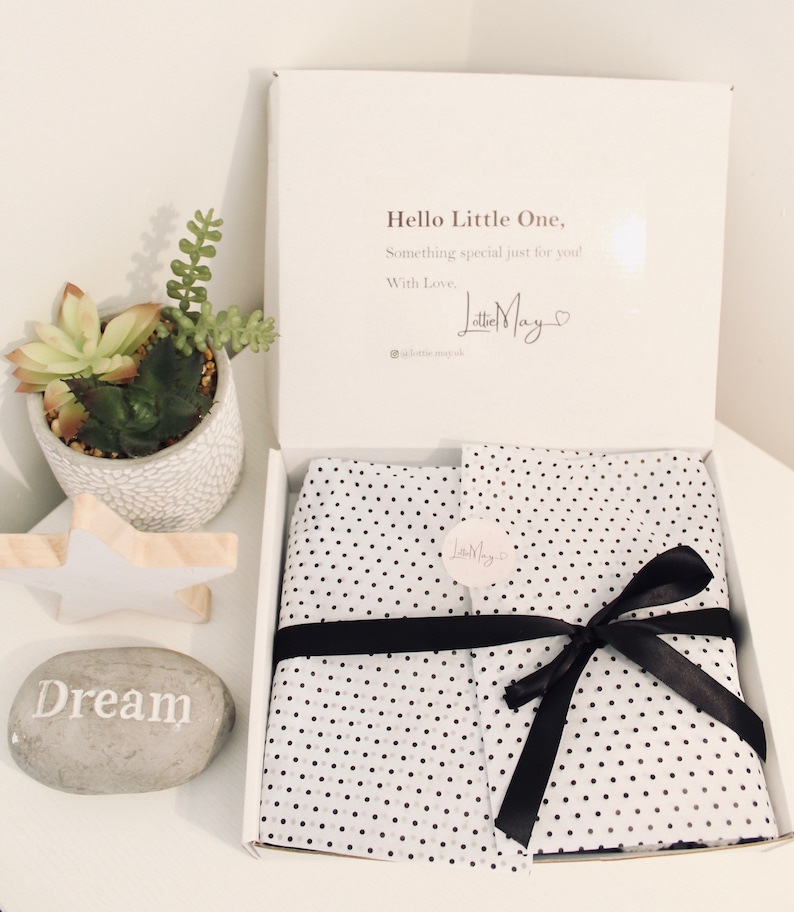 May include: A white gift box containing items wrapped in white tissue paper with black polka dots, secured by a black ribbon and a circular sticker. The inside lid features the text: Hello Little One, Something special just for you! With Love, Lottie May.