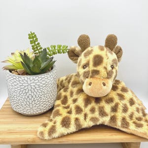 May include: A plush giraffe security blanket featuring a soft, tan and brown spotted fabric with a stuffed giraffe head and small horns. The giraffe has dark eyes and a light tan muzzle. The plush toy is positioned next to a small succulent plant.