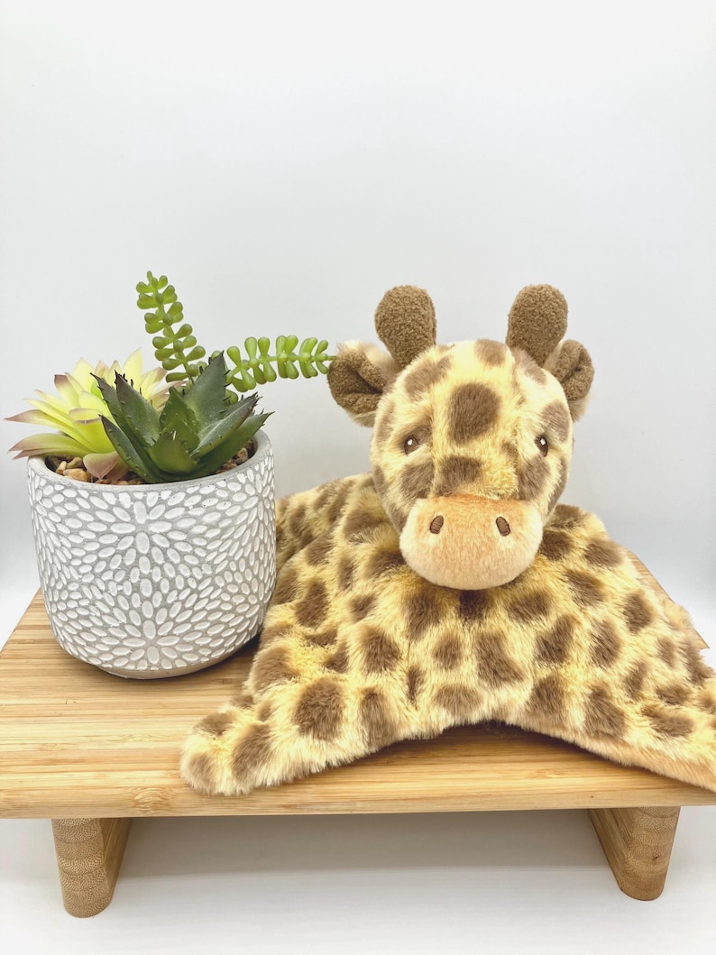 May include: A plush giraffe security blanket featuring a soft, tan and brown spotted fabric with a stuffed giraffe head and small horns. The giraffe has dark eyes and a light tan muzzle. The plush toy is positioned next to a small succulent plant.