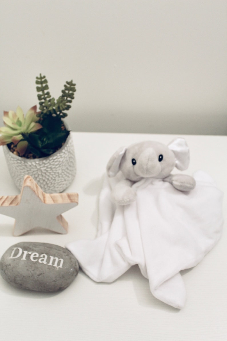 May include: A plush grey elephant security blanket with a white fabric body, a grey stone with the word Dream printed in white, a wooden star-shaped ornament, and a small green succulent plant in a textured grey pot.