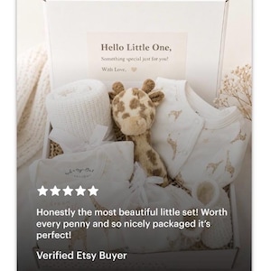 May include: A white gift box containing a baby gift set with a cream-coloured knit blanket, a plush giraffe toy, a white long-sleeve onesie with a gold giraffe pattern, and white booties. The box lid features the text: Hello Little One, Something special just for you! With Love.