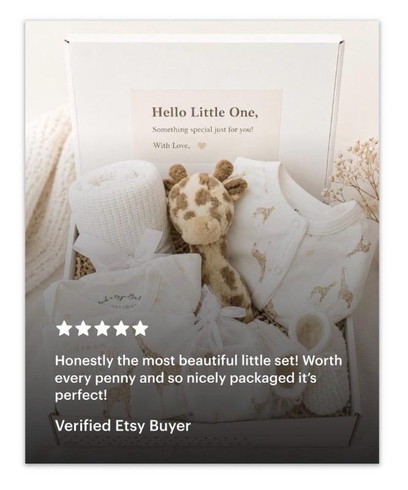 May include: A white gift box containing a baby gift set with a cream-coloured knit blanket, a plush giraffe toy, a white long-sleeve onesie with a gold giraffe pattern, and white booties. The box lid features the text: Hello Little One, Something special just for you! With Love.