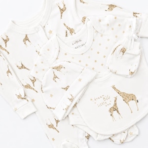 May include: A white baby clothing set featuring a long-sleeve footed sleepsuit, a short-sleeve bodysuit, a beanie, and mittens. The items are decorated with a gold giraffe pattern and stars. The bodysuit includes the text dream BIG little one.