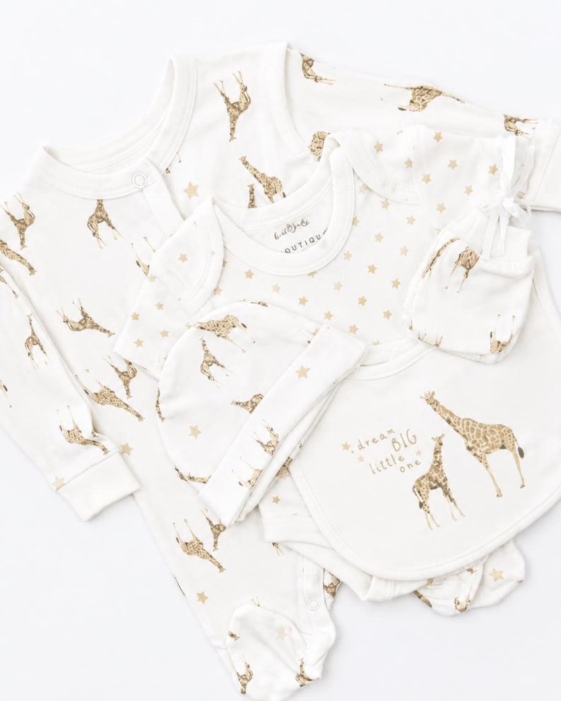 May include: A white baby clothing set featuring a long-sleeve footed sleepsuit, a short-sleeve bodysuit, a beanie, and mittens. The items are decorated with a gold giraffe pattern and stars. The bodysuit includes the text dream BIG little one.