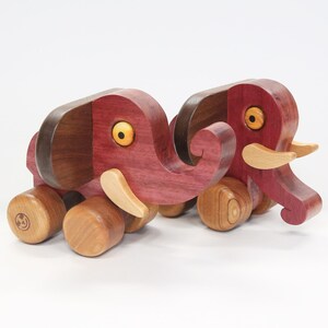 Op de afbeelding: Twee houten speelgoed olifanten met wielen. De olifanten zijn geschilderd in tinten paars en bruin. Ze hebben gele ogen en houten slurfjes.