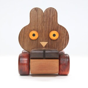 Op de afbeelding: Een houten speelgoedkonijn met een bruin lichaam, oranje ogen en een driehoekige neus. Het konijn heeft een rechthoekig lichaam en rode wielen. Het speelgoed is gemaakt van verschillende houttinten.