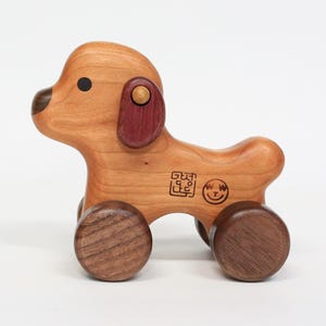 Puede incluir: Un perro de juguete de madera con ruedas marrones y una oreja roja. El perro tiene un ojo negro y una pequeña sonrisa tallada en su cara. El juguete está marcado con un pequeño cuadrado y una cara sonriente.