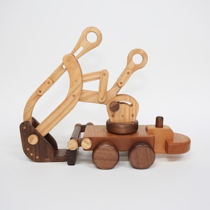 Op de afbeelding: Houten speelgoedgraafmachine in bruintinten. De graafmachine heeft een beweegbare arm en bak, gemonteerd op een vrachtwagen met vier wielen. Het speelgoed is gemaakt van verschillende houttinten en heeft een eenvoudig, klassiek ontwerp.