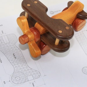 May include: A wooden toy airplane with brown and yellow wings, a brown body, and a propeller. The airplane is sitting on a white piece of paper with a blueprint design.