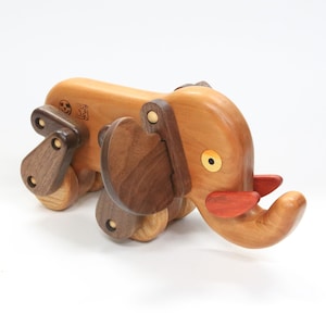 May include: A wooden toy elephant with a light brown body, dark brown legs and ears, and red accents on the trunk. The elephant has a round yellow eye and is designed with articulated joints. The toy is on a white background.