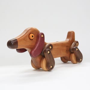 Puede incluir: Un juguete de madera en forma de dachshund con detalles en madera marrón, cerezo y morado. El juguete tiene una cabeza giratoria, patas móviles y cuatro ruedas. El juguete está hecho por "The Woodchuck Workshop".