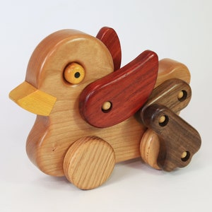 Può includere: Un'anatra giocattolo di legno con un becco giallo, ruote marroni e ali rosse e marroni. L'anatra è fatta di diversi tipi di legno, il che le conferisce un aspetto unico.