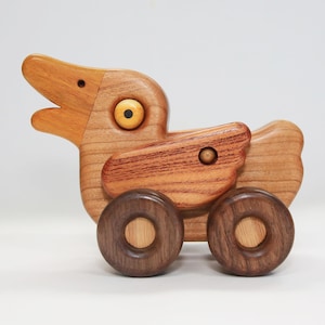 May include: A wooden toy duck with brown wheels and yellow eyes. The duck is made of light and dark wood and has a carved beak.