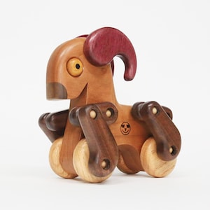Op de afbeelding: Een houten speelgoedram met een lichtbruin lichaam, donkerbruine poten en bordeauxrode hoorns. De ram heeft een lachend gezicht en is gemonteerd op houten wielen. Een klein logo is zichtbaar aan de zijkant.