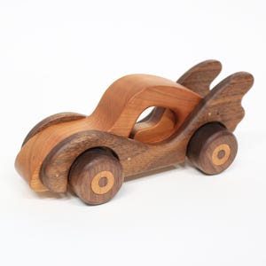 Può includere: Un'auto giocattolo in legno dal design aerodinamico. L'auto è realizzata con due tipi di legno, con un legno più chiaro per la carrozzeria e un legno più scuro per le ruote e gli accenti. L'auto ha un'esclusiva caratteristica a forma di ala sul retro.