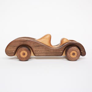 以下が含まれることがあります： クラシックなロードスターデザインの手作り木製おもちゃの車。車は、濃い茶色のボディに薄茶色のアクセントと木製の車輪が特徴です。おもちゃの車の長さは約25cmです。