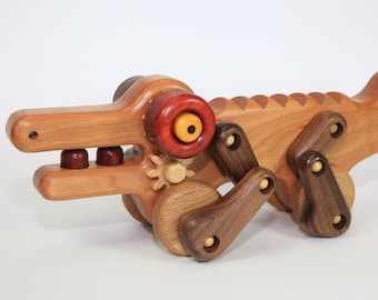 May include: A wooden alligator toy with articulated legs and a gear-driven jaw. The toy is crafted from various wood tones, including light and dark browns, with a red eye and yellow iris. The alligator's body features a ridged spine.
