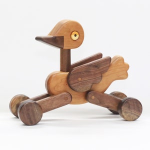 May include: A wooden bird toy with a light brown body, dark brown wings, and wheels. The bird has a long beak and a round, yellow eye. The toy is made of various wood pieces and is designed to roll.