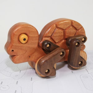 May include: A wooden toy turtle with a brown shell and a light brown head. The turtle has a yellow eye and is on wheels. The wheels are made of dark wood and have a light wood centre.