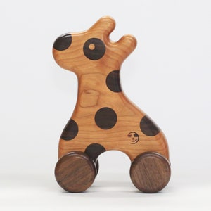 May include: Wooden giraffe pull toy with dark brown spots and wheels. The giraffe is made of light brown wood with a natural finish. The wheels are a darker brown. A small logo is visible on the side. A classic toy for toddlers.