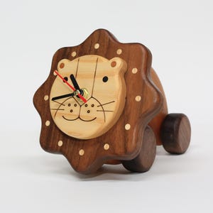May include: A wooden lion-shaped clock with a light brown face and dark brown mane and body. The clock face features black hands and a red second hand. The clock is mounted on wheels, giving it a toy-like appearance.
