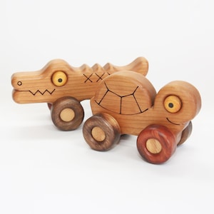 May include: Two wooden toy cars, one shaped like an alligator and the other shaped like a turtle. Both cars have yellow eyes and brown wheels.