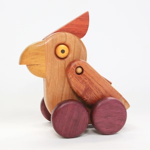 May include: A wooden toy bird with a red crest, yellow eye, and brown body. The bird has a beak and wing, and rolls on four purple wheels. A handcrafted wooden toy.
