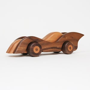 May include: A wooden toy race car with a streamlined design. The car is crafted from two tones of wood, with darker wood used for the body and wheels, and lighter wood for the top. The wheels have a circular design. A classic toy.