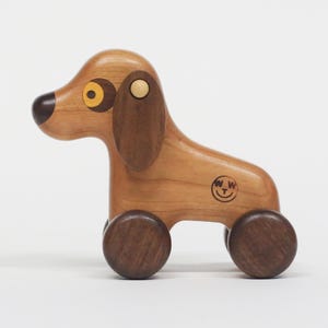 May include: A wooden pull-along toy dog with a light brown body, dark brown ears, nose, and wheels. The dog has yellow eyes and a small logo on its side. The toy is designed with rounded edges and a simple, classic aesthetic.