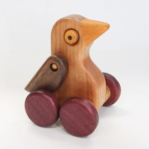 May include: A wooden toy bird with a brown wing, yellow eye, and purple wheels. The bird is made of cherry wood and has a natural finish.