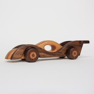 May include: A wooden toy car with a streamlined design. The car is made of various wood tones, including light and dark brown. It features a circular opening in the center and wooden wheels. The car has a spoiler at the back and is approximately 30 cm long.