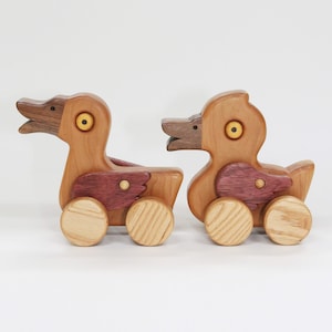 May include: Two wooden duck toys with wheels. The ducks are made of light brown wood with darker brown heads and purple-red wings. The eyes are yellow. The wheels are light brown with a striped design. The toys are on a white background.