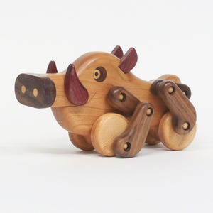 May include: A wooden toy warthog with a light brown body, dark brown snout, and purple accents. The toy has wheels and articulated legs, giving it a playful appearance. The eyes are a dark brown color.