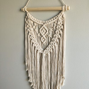 May include: A beige macrame wall hanging with a wooden dowel at the top. The hanging features a geometric pattern with fringe at the bottom.
