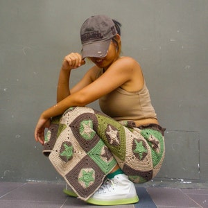 May include: A person wearing a brown baseball cap with the text "Good Luck" on it, a beige tank top, and a green and brown crocheted skirt. The person is sitting on the ground with their legs crossed.