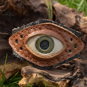 May include: A brown leather bracelet with a large, white eye design. The eye has a green iris and a black pupil.