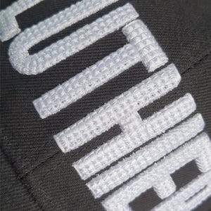 May include: Close-up of a black fabric with white embroidered text. The white text is thick and textured, with a woven appearance. The letters are large and bold, creating a striking contrast against the dark background.
