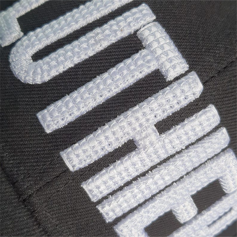 May include: Close-up of a black fabric with white embroidered text. The white text is thick and textured, with a woven appearance. The letters are large and bold, creating a striking contrast against the dark background.