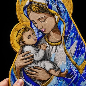 Könnte beinhalten: Ein detaillierter gestickter Aufnäher mit einer stilisierten Darstellung einer Figur, die ein Kind hält. Das Design verwendet leuchtend blaue, weiße und goldene Fäden mit einem markanten goldenen Heiligenschein vor schwarzem Hintergrund.