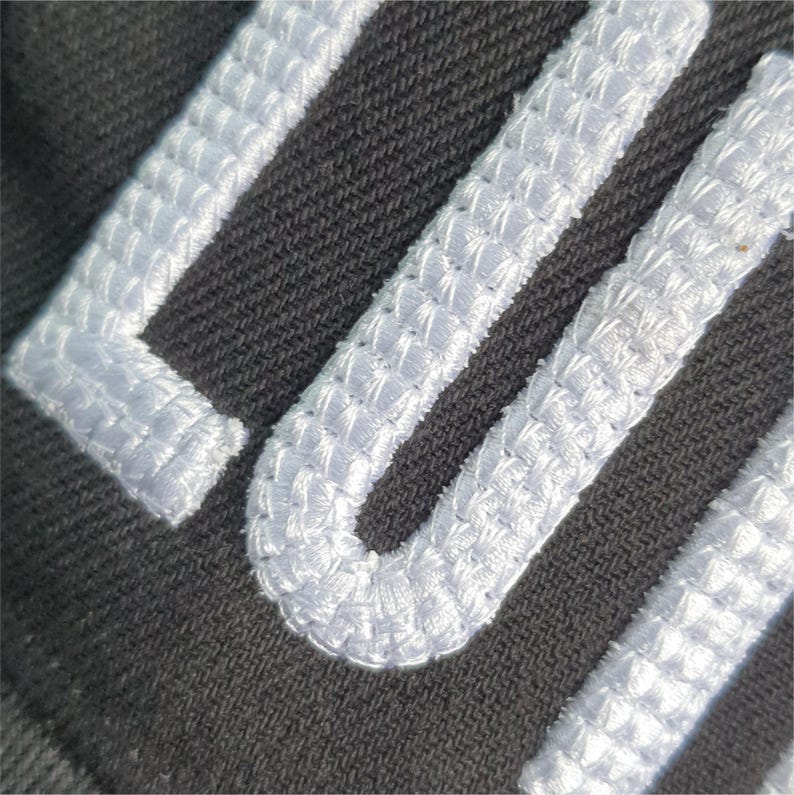 May include: Close-up of dark gray fabric with white embroidered lettering. The white embroidery has a textured, raised appearance, creating a 3D effect against the darker background. The stitching is detailed and precise.
