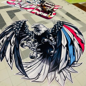 Peut inclure: Un aigle chauve noir et blanc avec ses ailes d&eacute;ploy&eacute;es. L'aigle tient un petit drapeau am&eacute;ricain dans ses serres. Les ailes de l'aigle sont d&eacute;cor&eacute;es des couleurs du drapeau am&eacute;ricain, rouge, blanc et bleu.