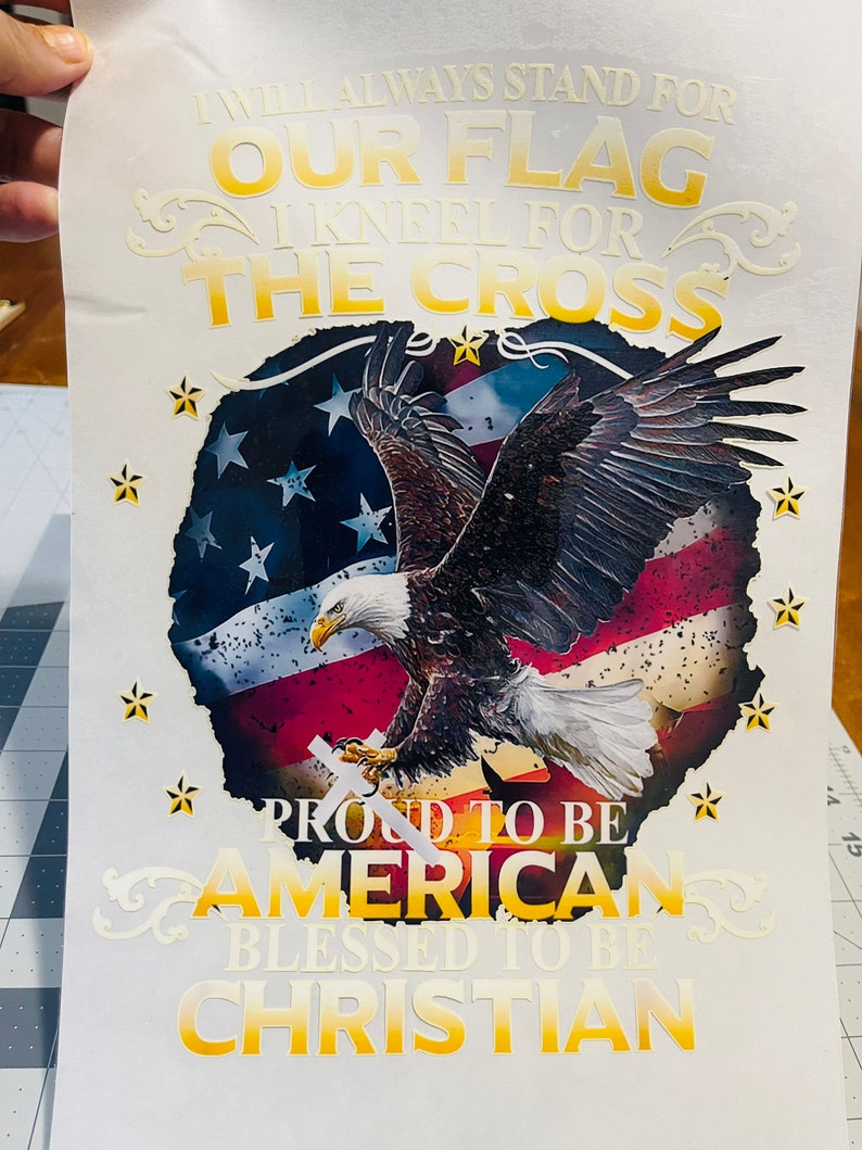 Peut inclure: Un fond blanc avec un design graphique or et blanc repr&eacute;sentant un aigle chauve am&eacute;ricain avec un drapeau am&eacute;ricain en arri&egrave;re-plan. Le texte dit "I will always stand for our flag I kneel for the cross Proud to be American Blessed to be Christian".