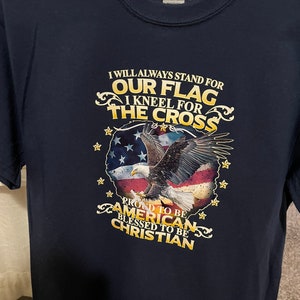 Peut inclure: Un t-shirt bleu marine avec un graphique d'un aigle volant au-dessus d'un drapeau am&eacute;ricain. Le texte "I will always stand for our flag I kneel for the cross Proud to be American Blessed to be Christian" est imprim&eacute; sur le t-shirt.