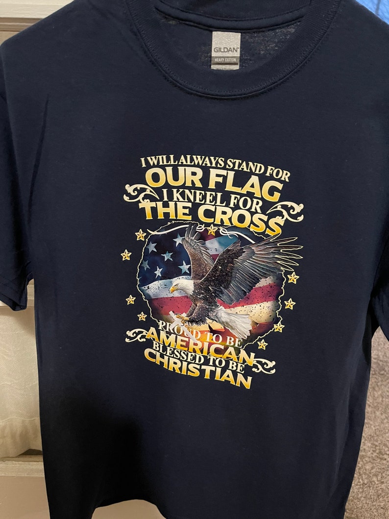 Peut inclure: Un t-shirt bleu marine avec un graphique d'un aigle volant au-dessus d'un drapeau am&eacute;ricain. Le texte "I will always stand for our flag I kneel for the cross Proud to be American Blessed to be Christian" est imprim&eacute; sur le t-shirt.